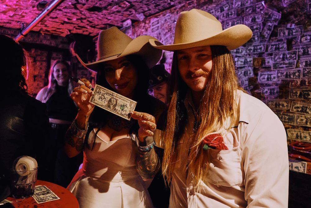 A wedding party celebrates in a dimly lit bar with dollar bills decorating the ceiling.