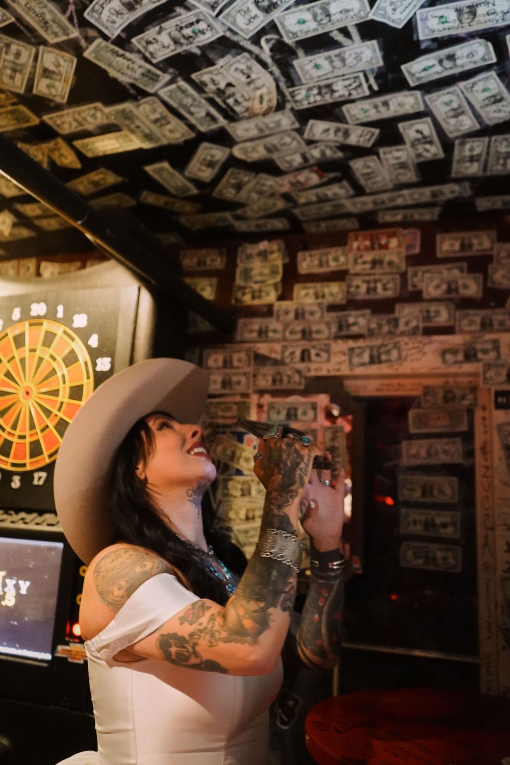 A bar interior with a dartboard visible and dollar bills covering the entire ceiling.