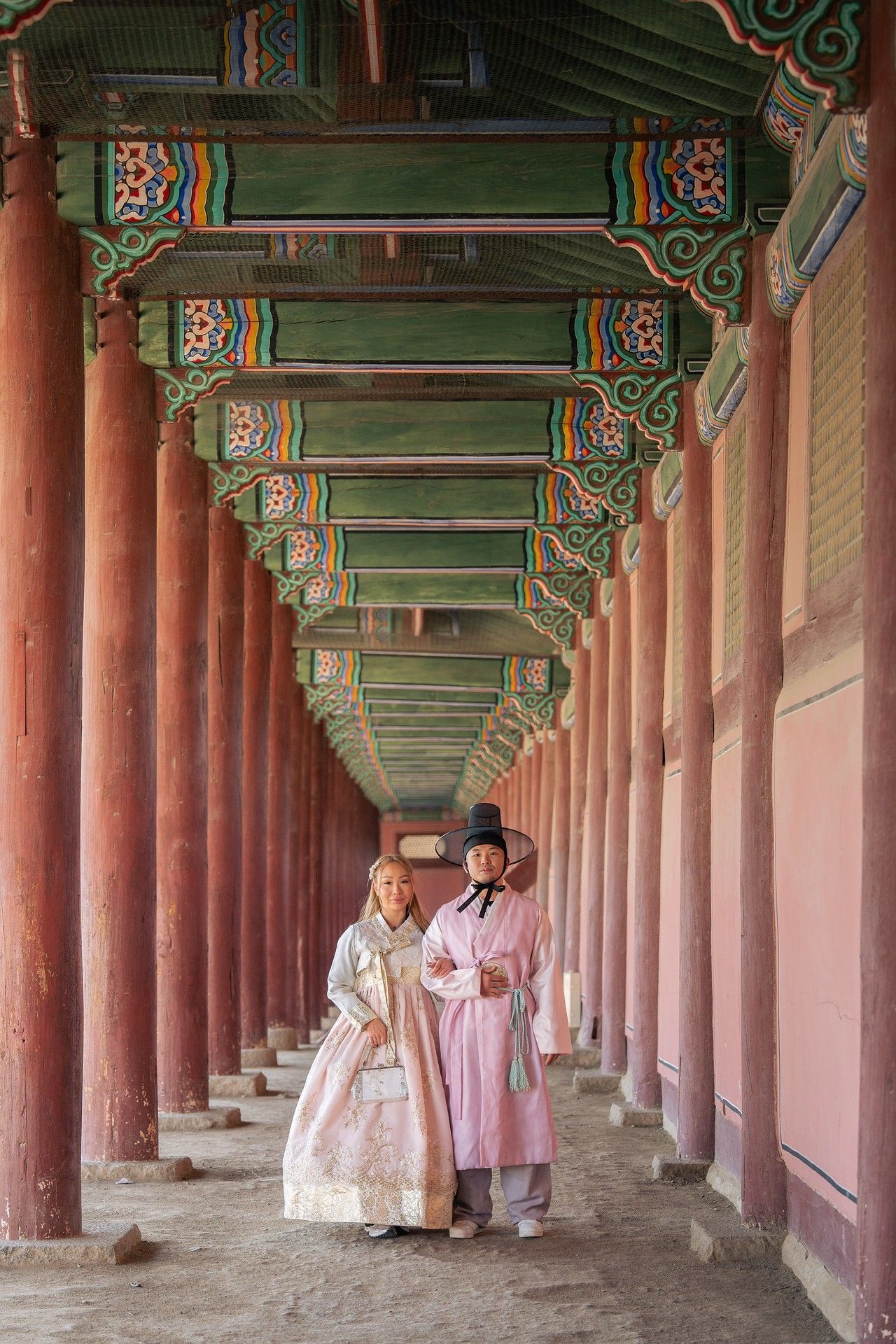 People in traditional Korean hanbok clothing walk through a long covered corridor with ornate red pillars at Gyeongbokgung Palace.