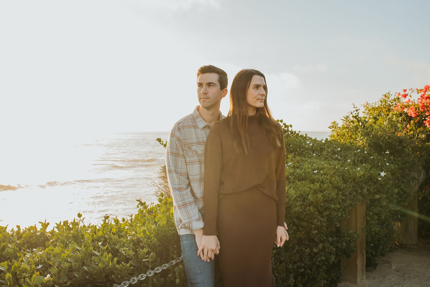 A couple stands along a scenic coastal path at sunset with green foliage and ocean views in the background at Windansea Beach.