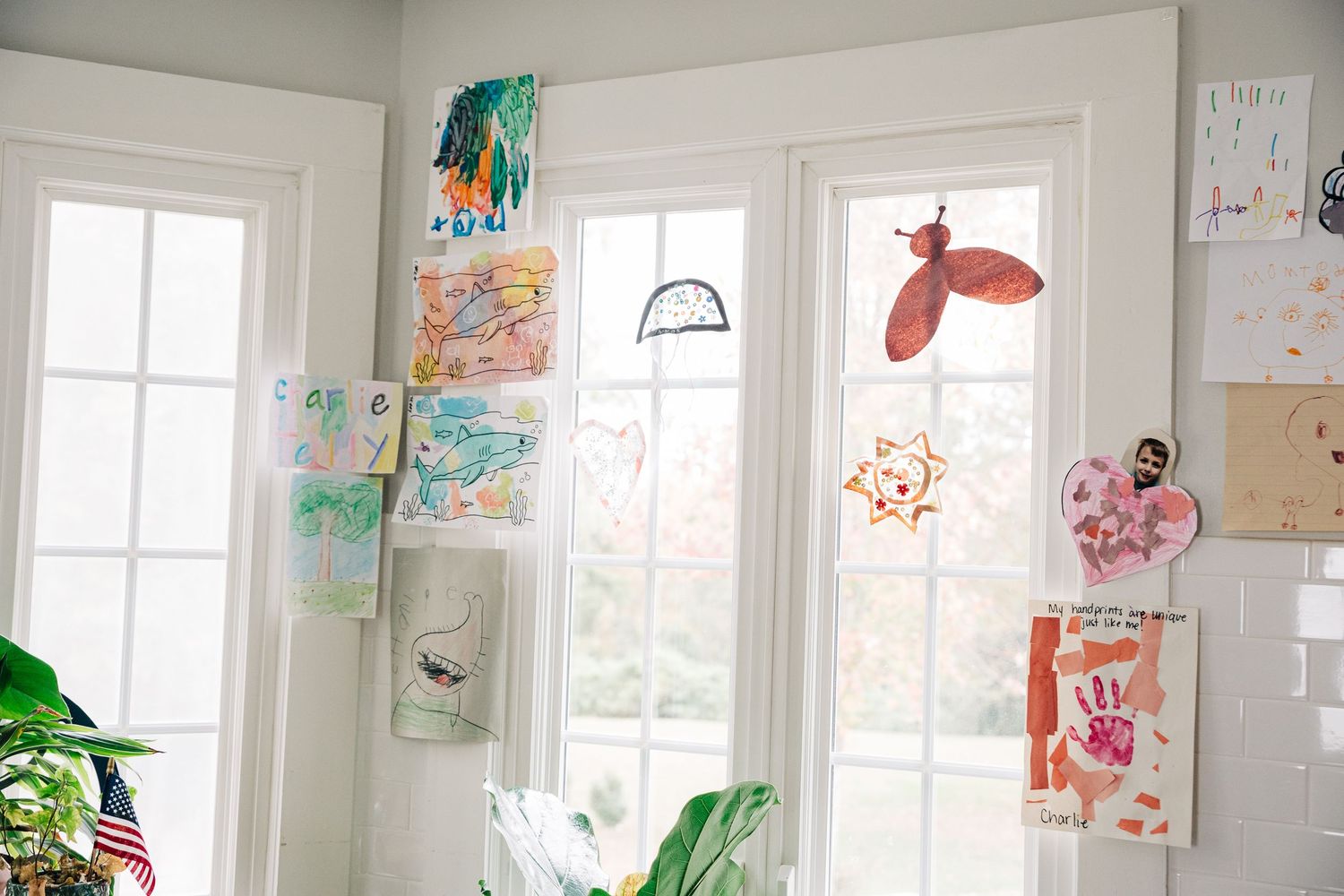 Children's artwork and drawings displayed on white walls near bright windows with houseplants in a sunny room.