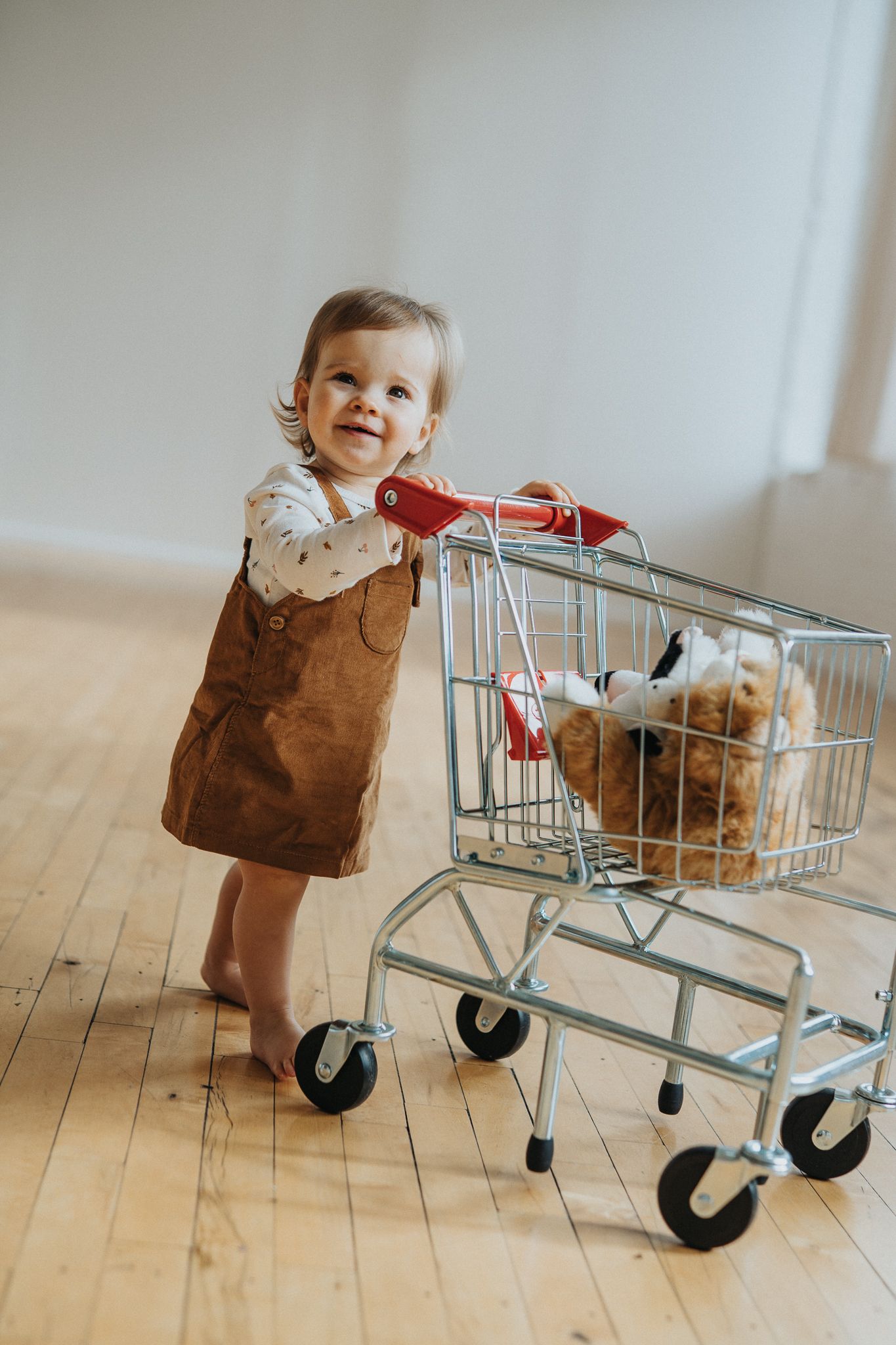 A small child pushes a shopping cart across a wooden floor while wearing a brown dress.