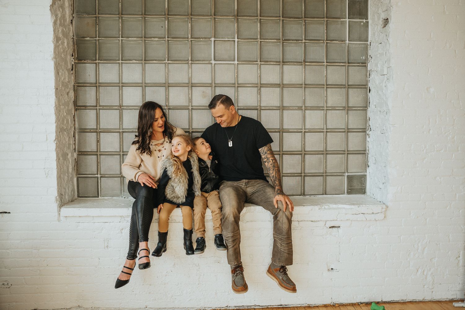 Three people sit together on a ledge against a white wall with a glass block window.