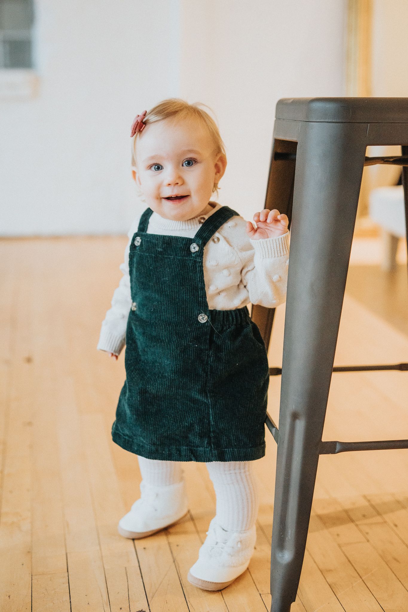 A toddler in a green corduroy jumper dress and white tights stands holding onto a chair on a hardwood floor.