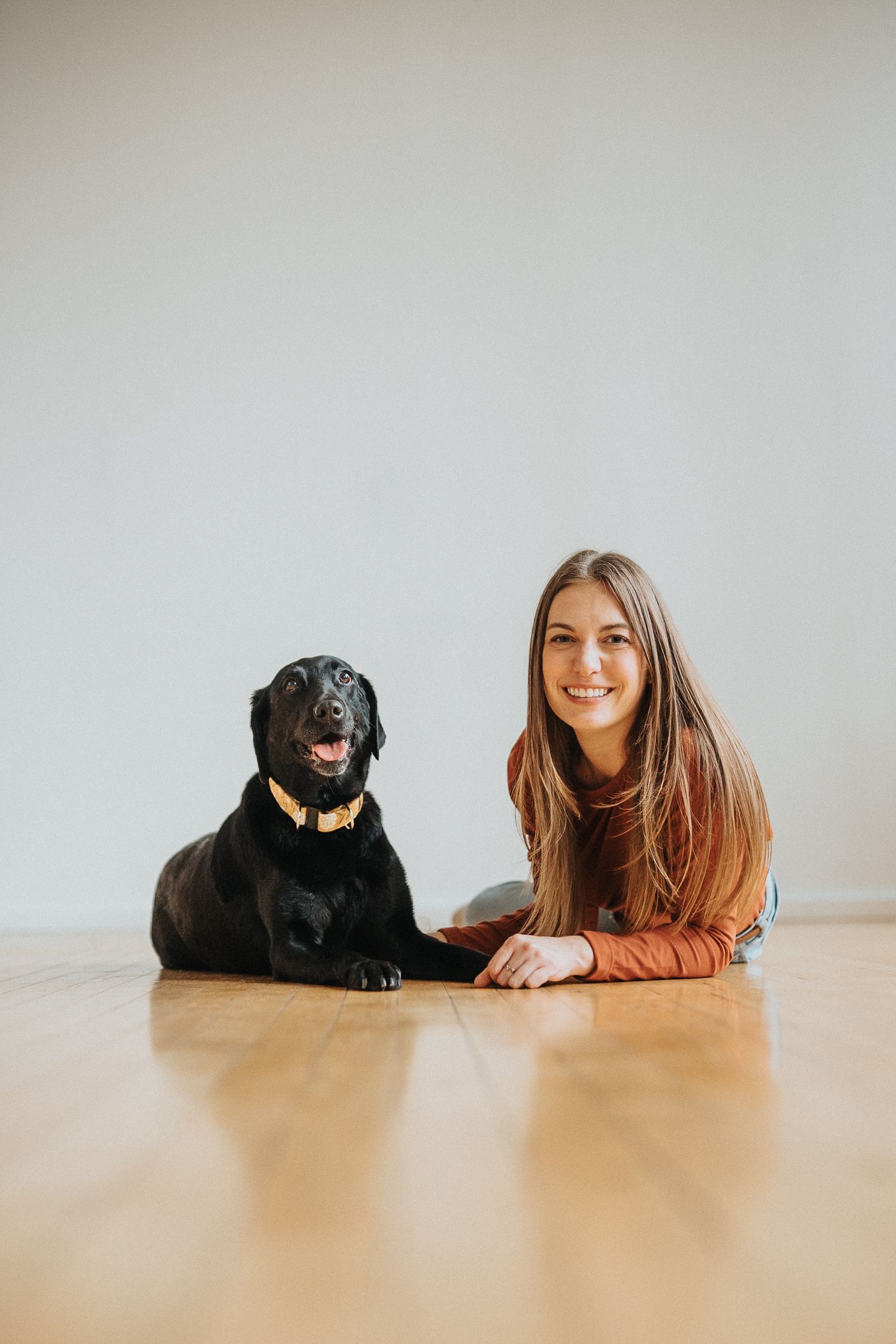 A person in a brown sweater lies on hardwood floor next to their black Labrador retriever companion.