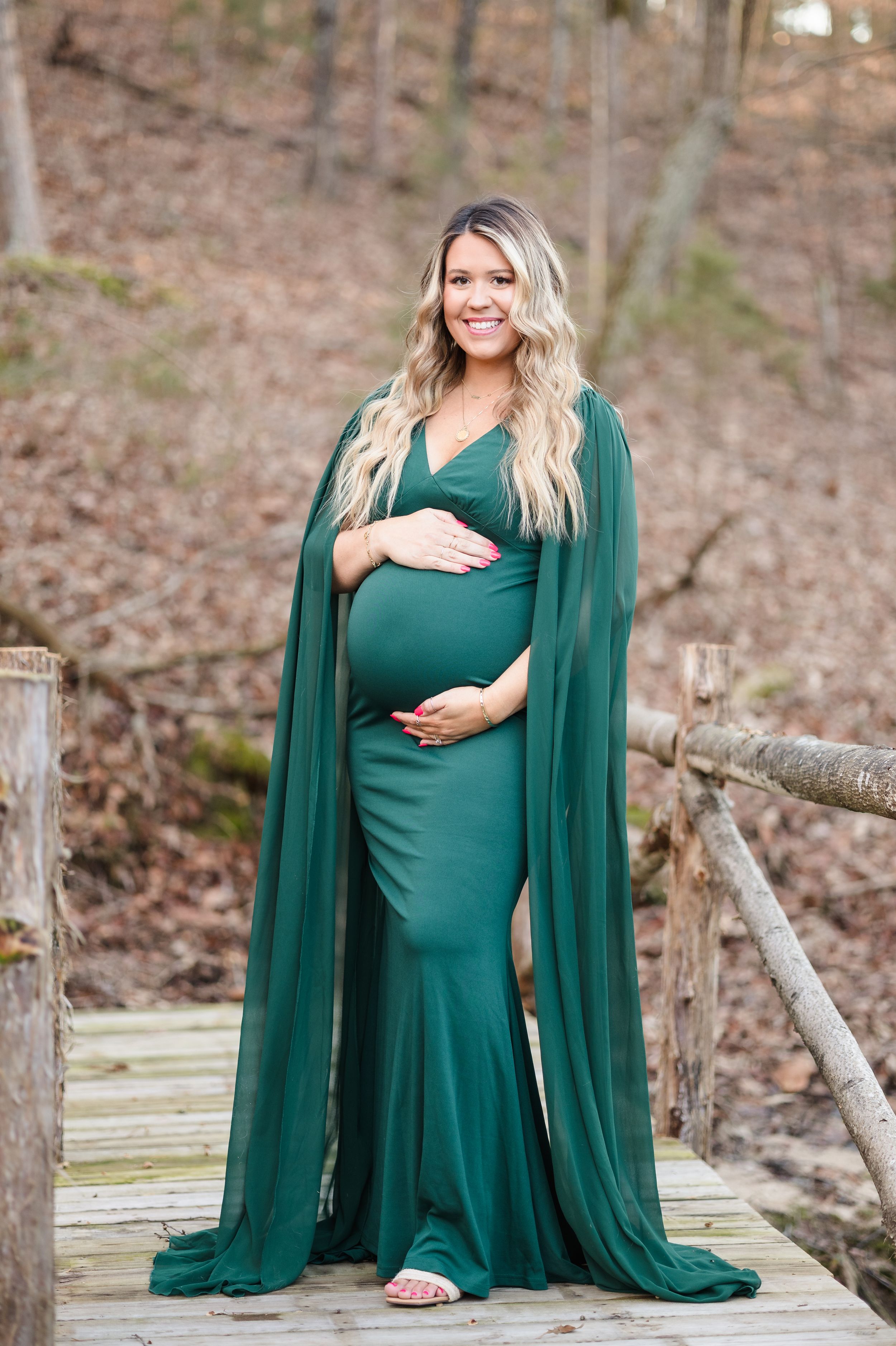 Pregnant woman in flowing emerald green gown and cape poses on wooden bridge in forest setting.