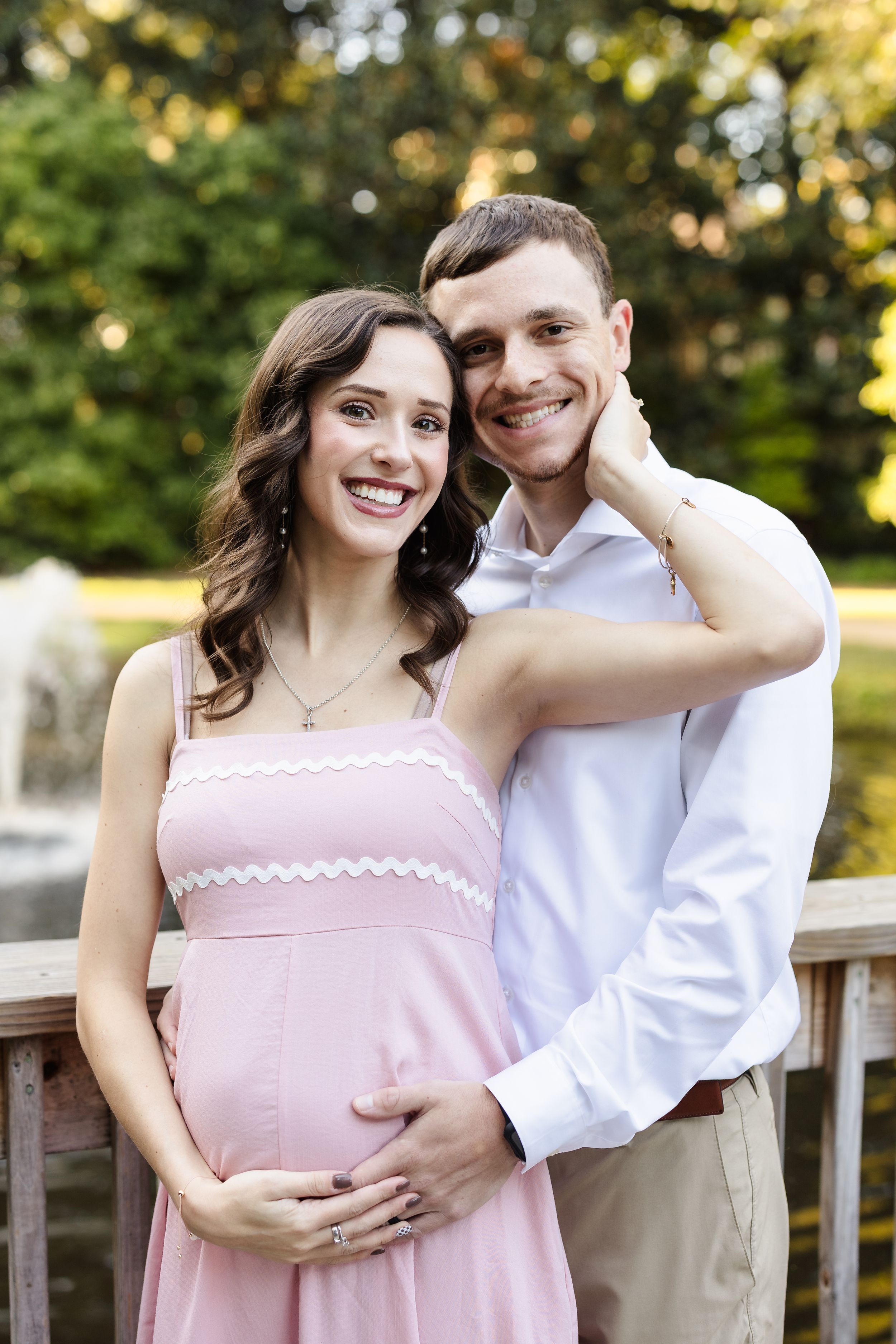 Couple in pink dress and white shirt posing together on wooden deck during sunset.