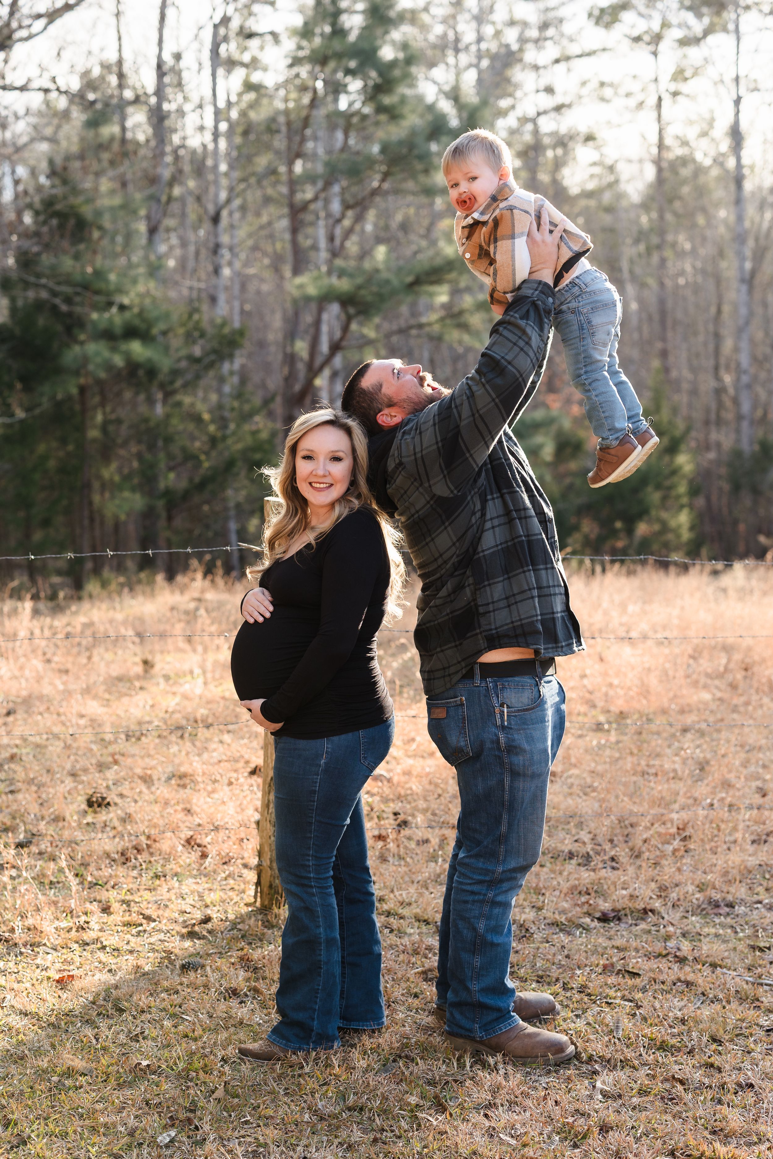 Family outdoor photo with pregnant woman in black while partner lifts child in autumn field.
