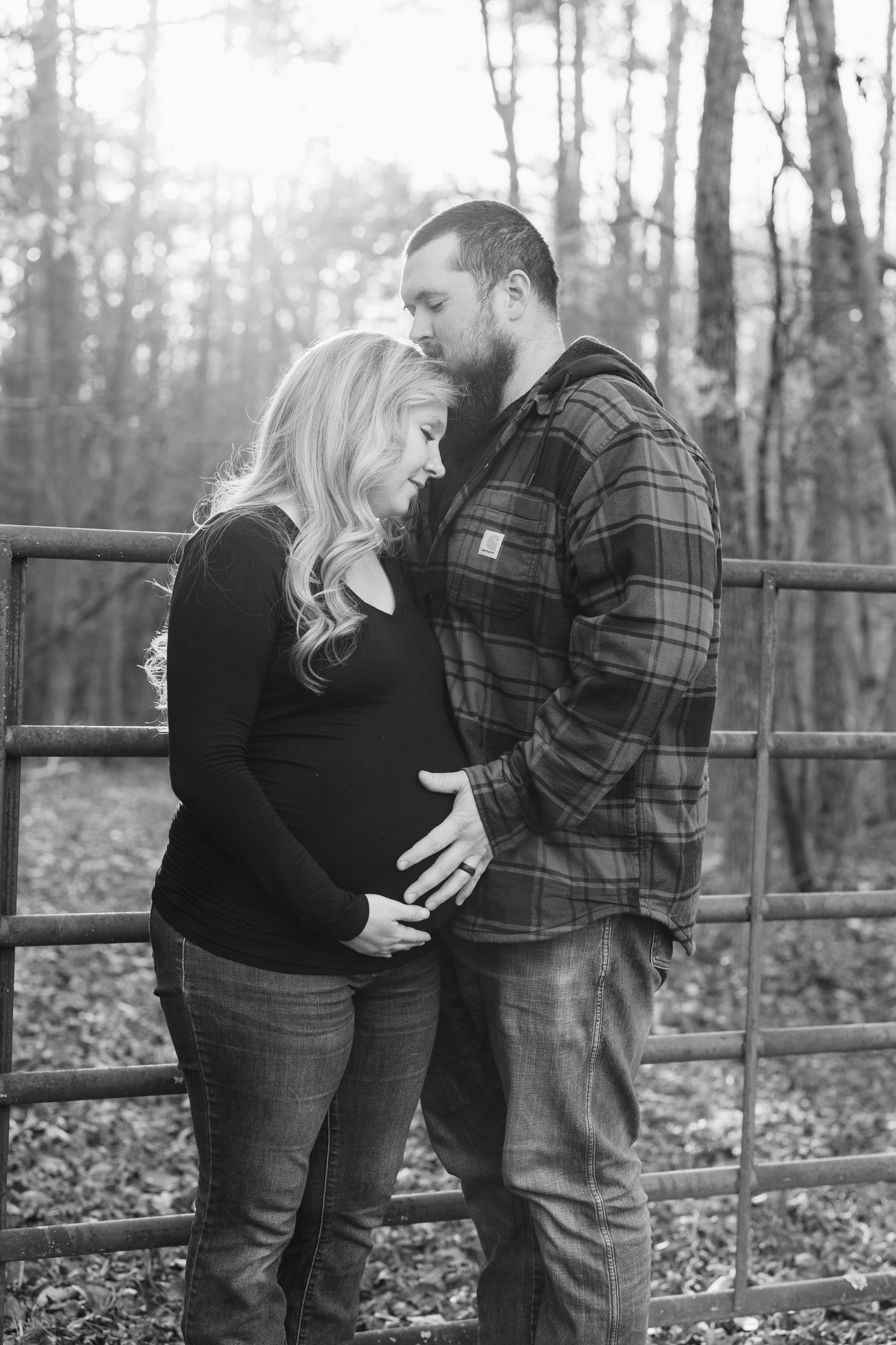 Black and white maternity photo of couple embracing by rustic fence in forest setting.