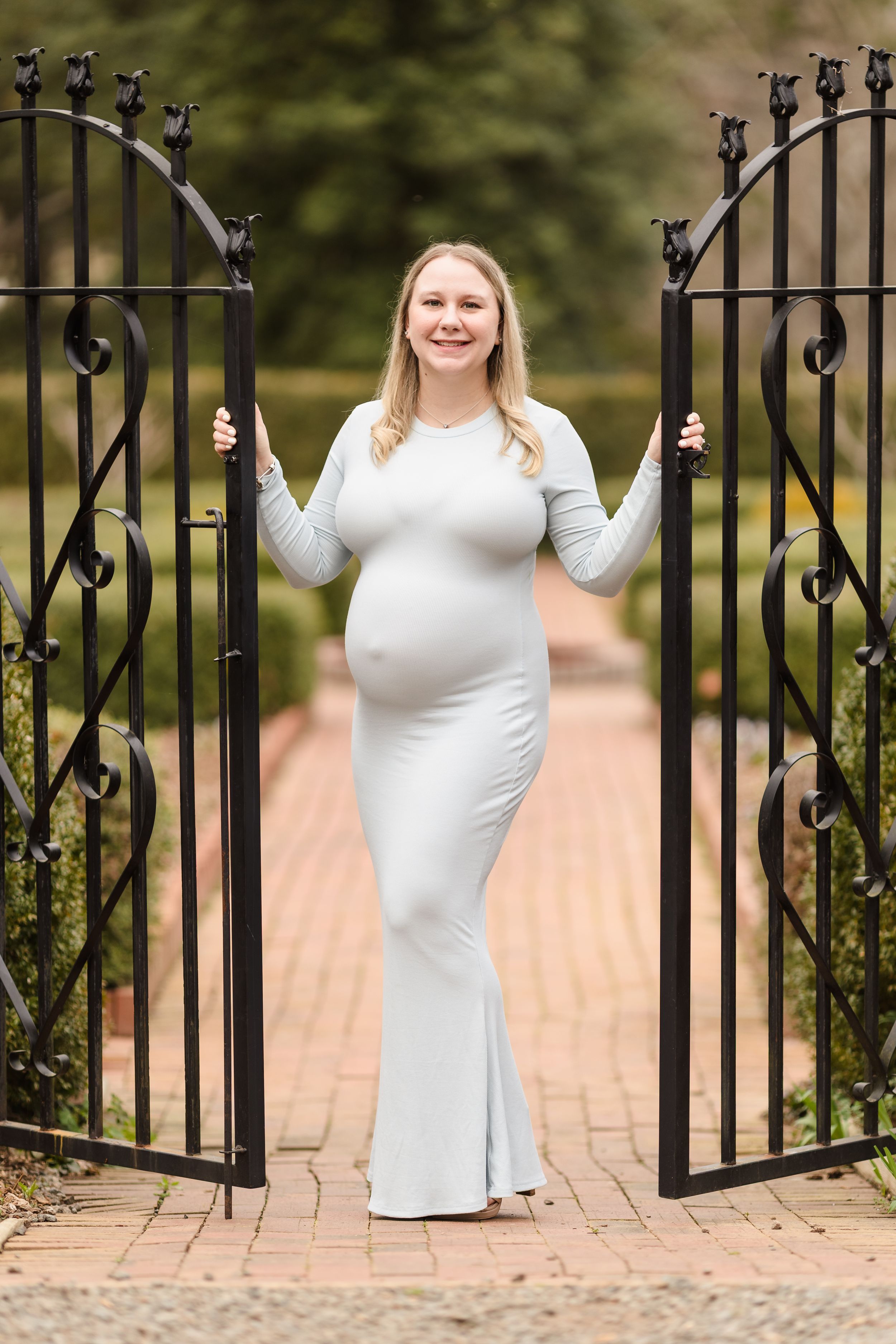 A person in a white dress poses elegantly by a decorative iron gate in an outdoor setting.