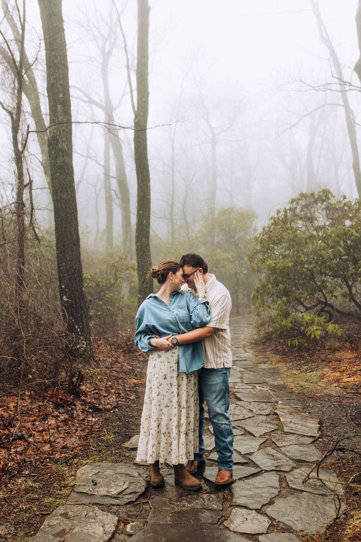 A romantic couple shares an intimate moment on a foggy stone path surrounded by misty bare trees.
