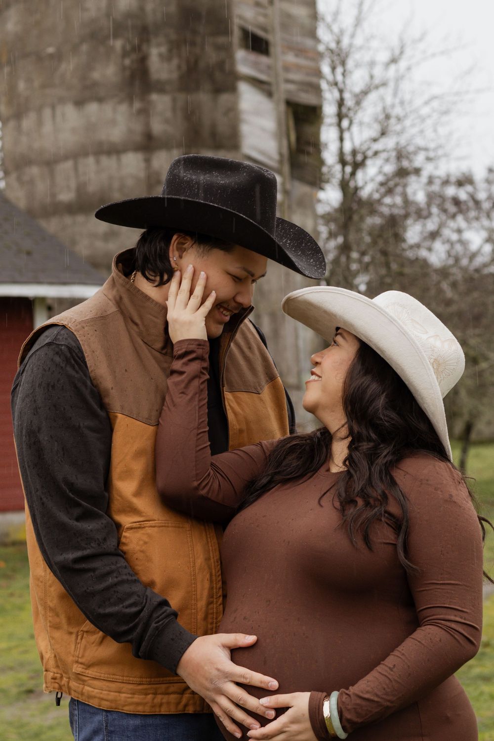 Expecting couple in cowboy hats sharing a loving glance near a rustic barn in the rain.
