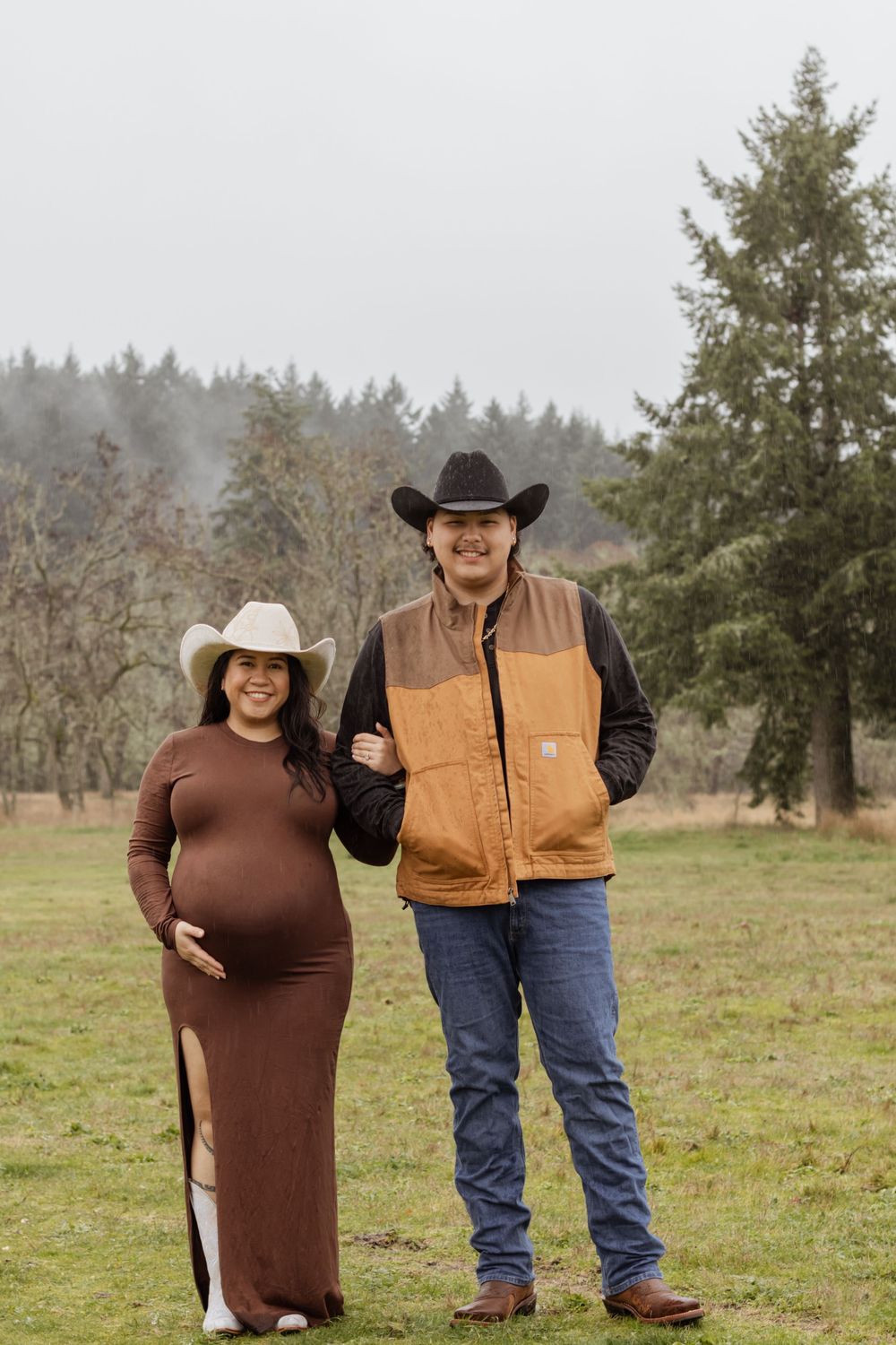 Smiling pregnant couple in cowboy hats posing in a misty green field on a farm.