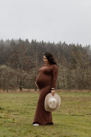 Pregnant woman in brown maxi dress holding cream cowboy hat in misty field.