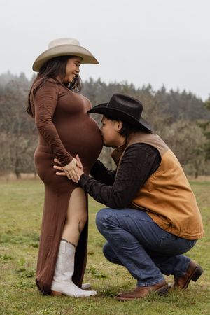Partner kissing pregnant belly while couple poses in cowboy hats outdoors.