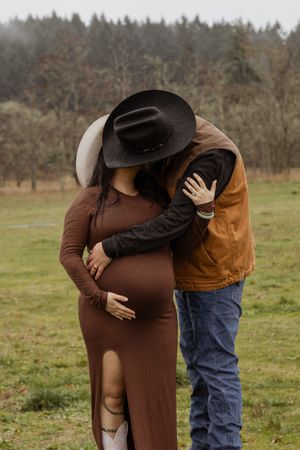 Couple in cowboy hats embracing tenderly in rustic field maternity photo.