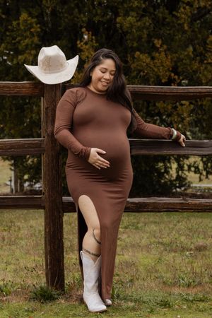 Smiling pregnant woman in brown dress and white boots leaning on wooden fence.