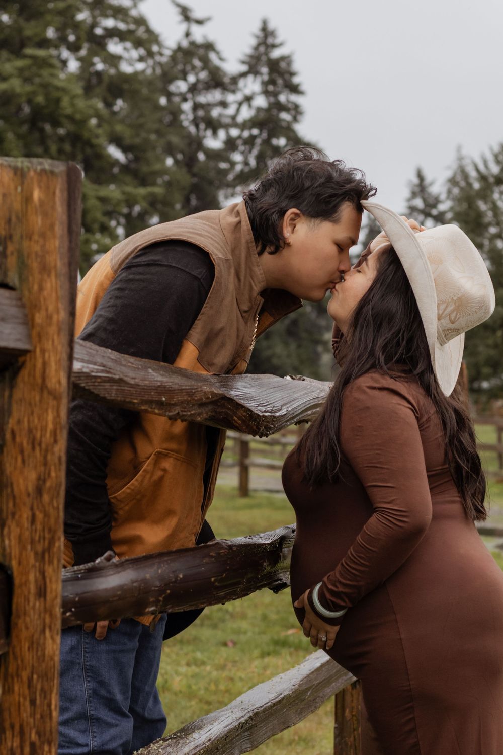 Couple sharing a kiss over rustic wooden fence during maternity photoshoot.