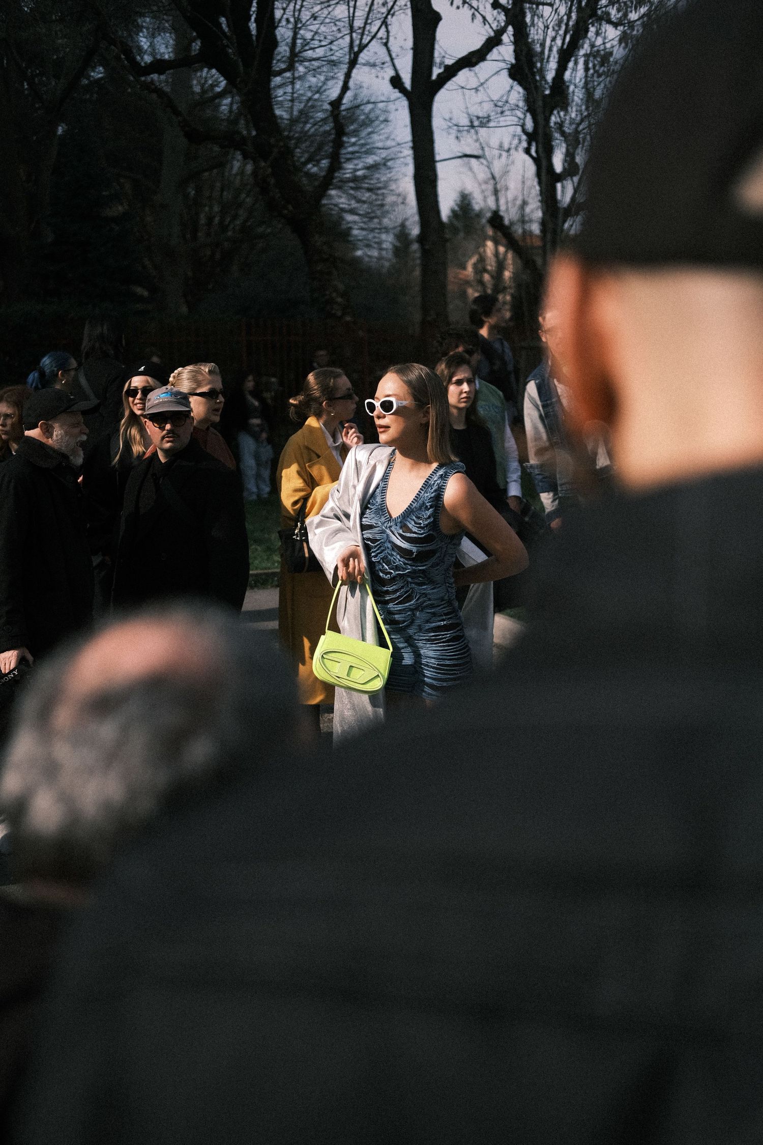 Group of people gathered outdoors in winter, some holding cameras and phones, against a backdrop of bare trees.
