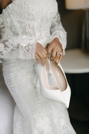 Bride in lace wedding dress holding white pointed-toe bridal heels with diamond engagement ring.