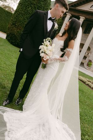 Bride in a lace wedding gown and long veil holding a white rose bouquet beside a groom in a black tuxedo on a manicured lawn.