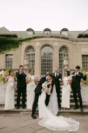 Bride and groom kiss on stone steps as bridesmaids and groomsmen cheer in front of a historic ivy-covered wedding venue.