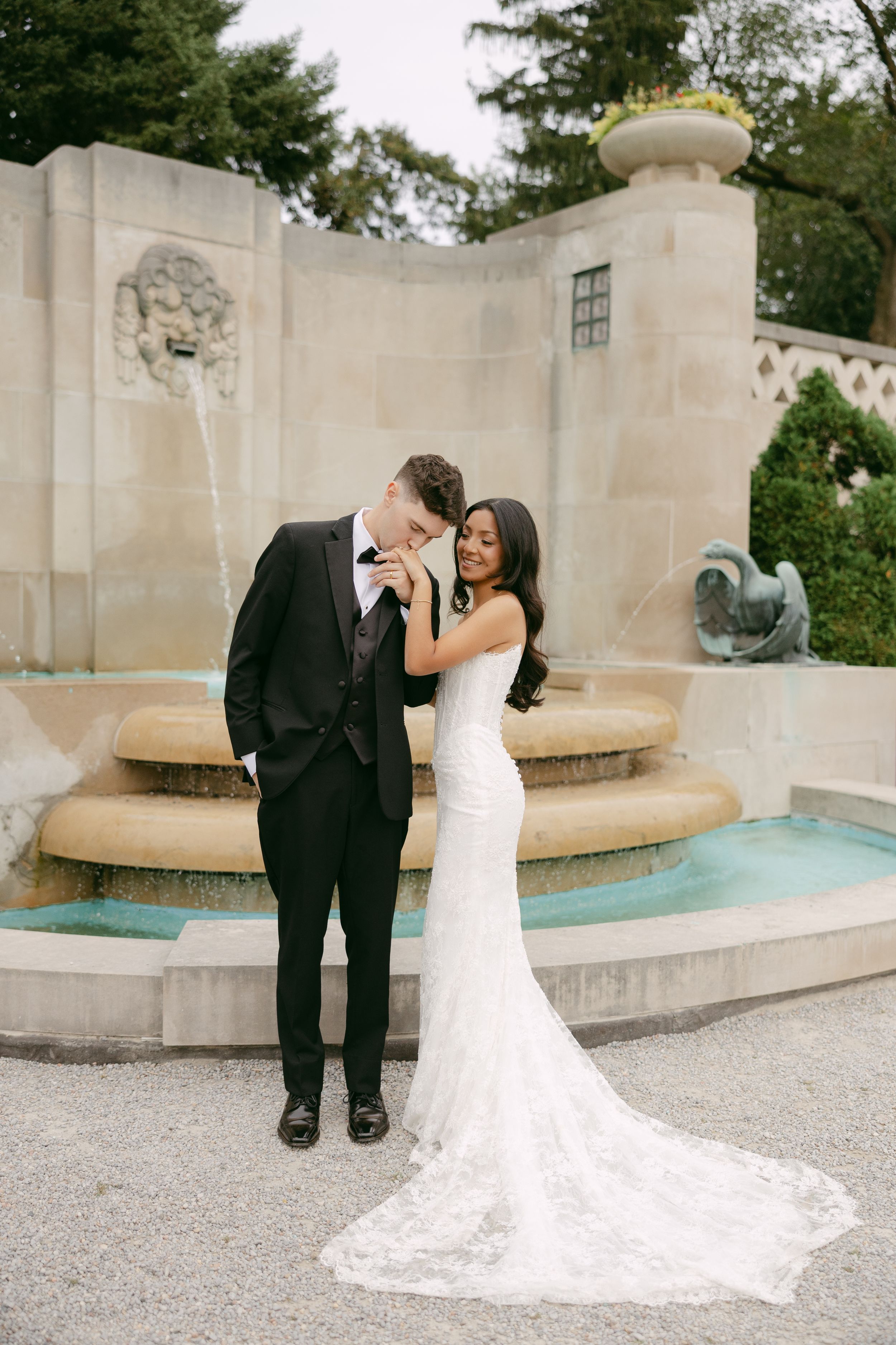 Bride in a lace mermaid wedding gown kisses the hand of her groom in a black tuxedo beside a tiered stone fountain.