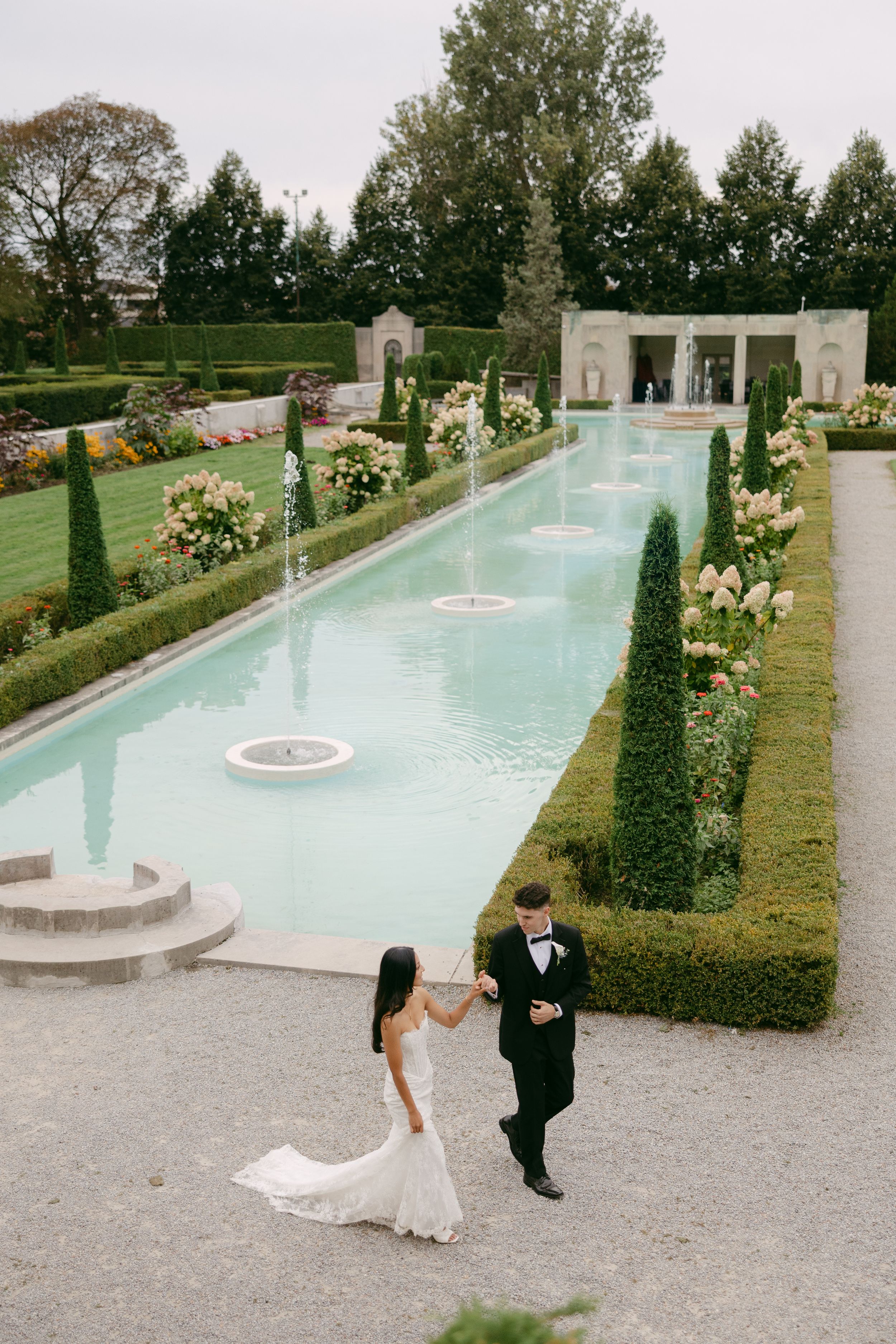Bride in a white gown and groom in a black tuxedo walk hand in hand beside a long reflecting pool in a formal garden.