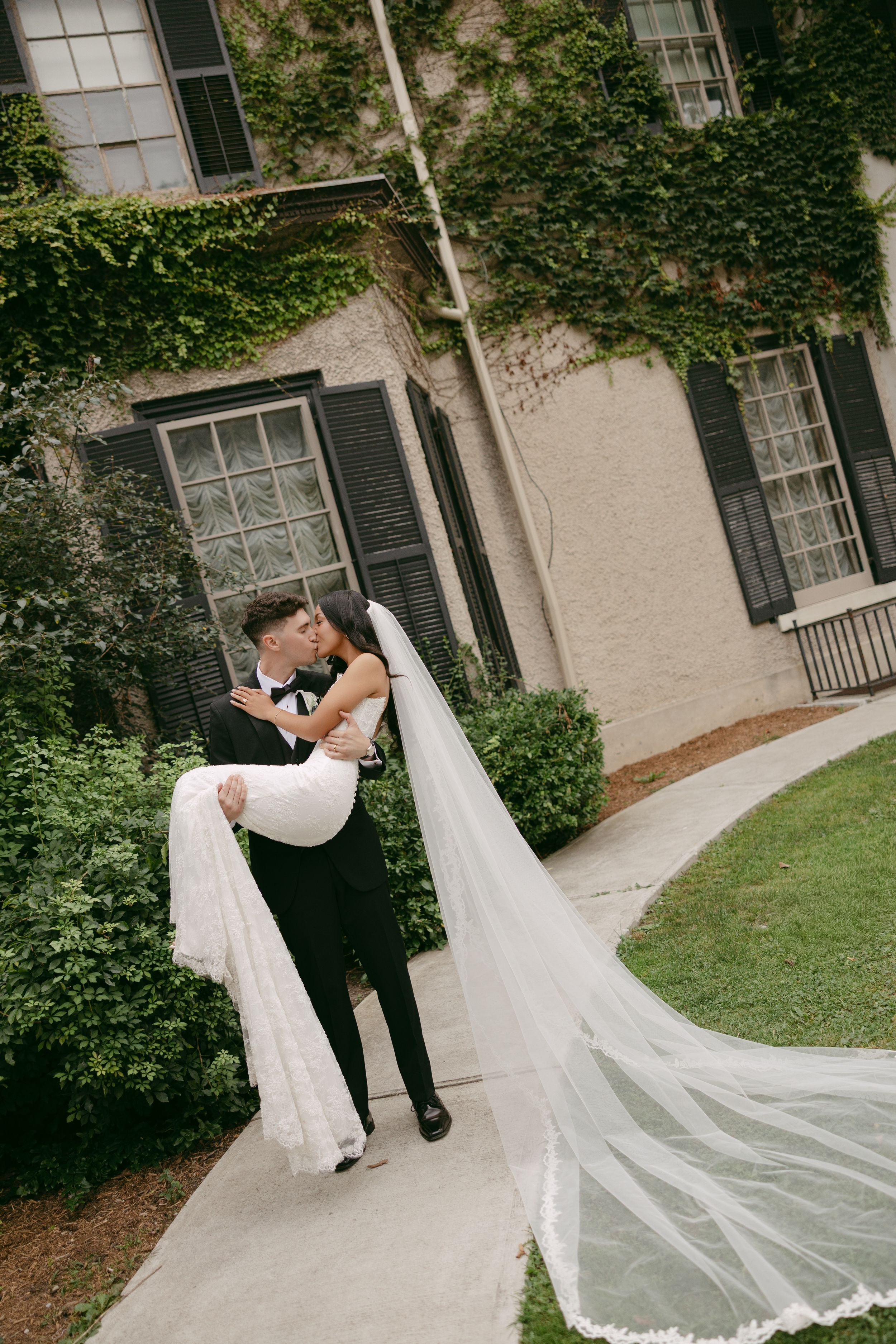 A groom in a black tuxedo cradles his bride in a white gown with a long lace-edged veil outside an ivy-covered stone building.