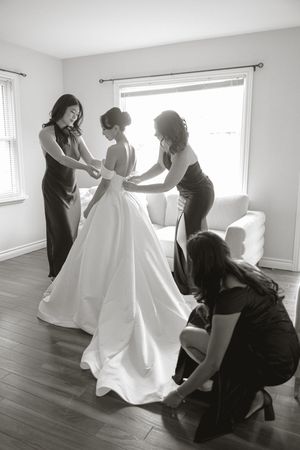 Three bridesmaids in dark dresses help a bride into her white ball gown beside a bright window in a black and white wedding photo.