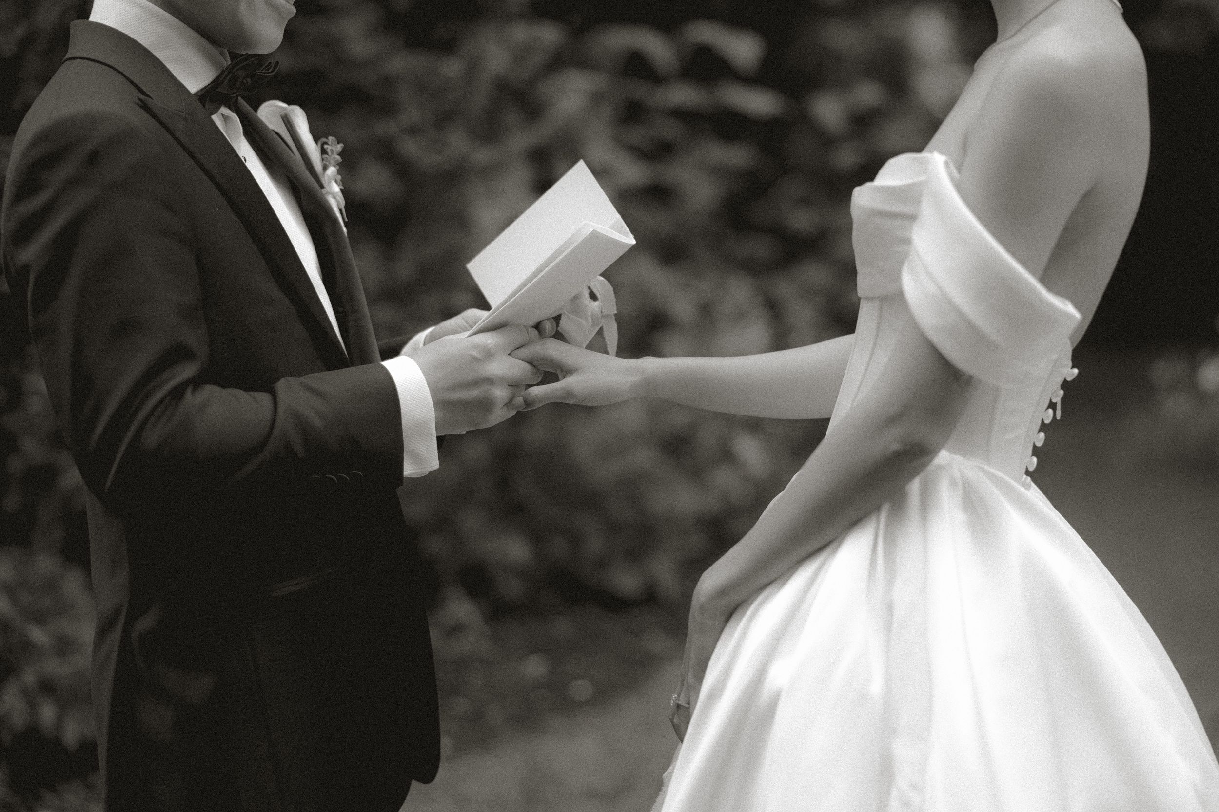 Groom in a dark suit reads his handwritten vows while holding the bride's hand during an outdoor wedding ceremony, black and white photo.