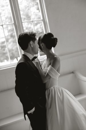 Black and white wedding photo of a bride in an off-shoulder gown and groom in a tuxedo sharing an intimate moment by a bright window.