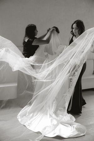 Two women help a bride adjust her flowing lace-trimmed veil and satin wedding gown in a black and white bridal preparation photo.