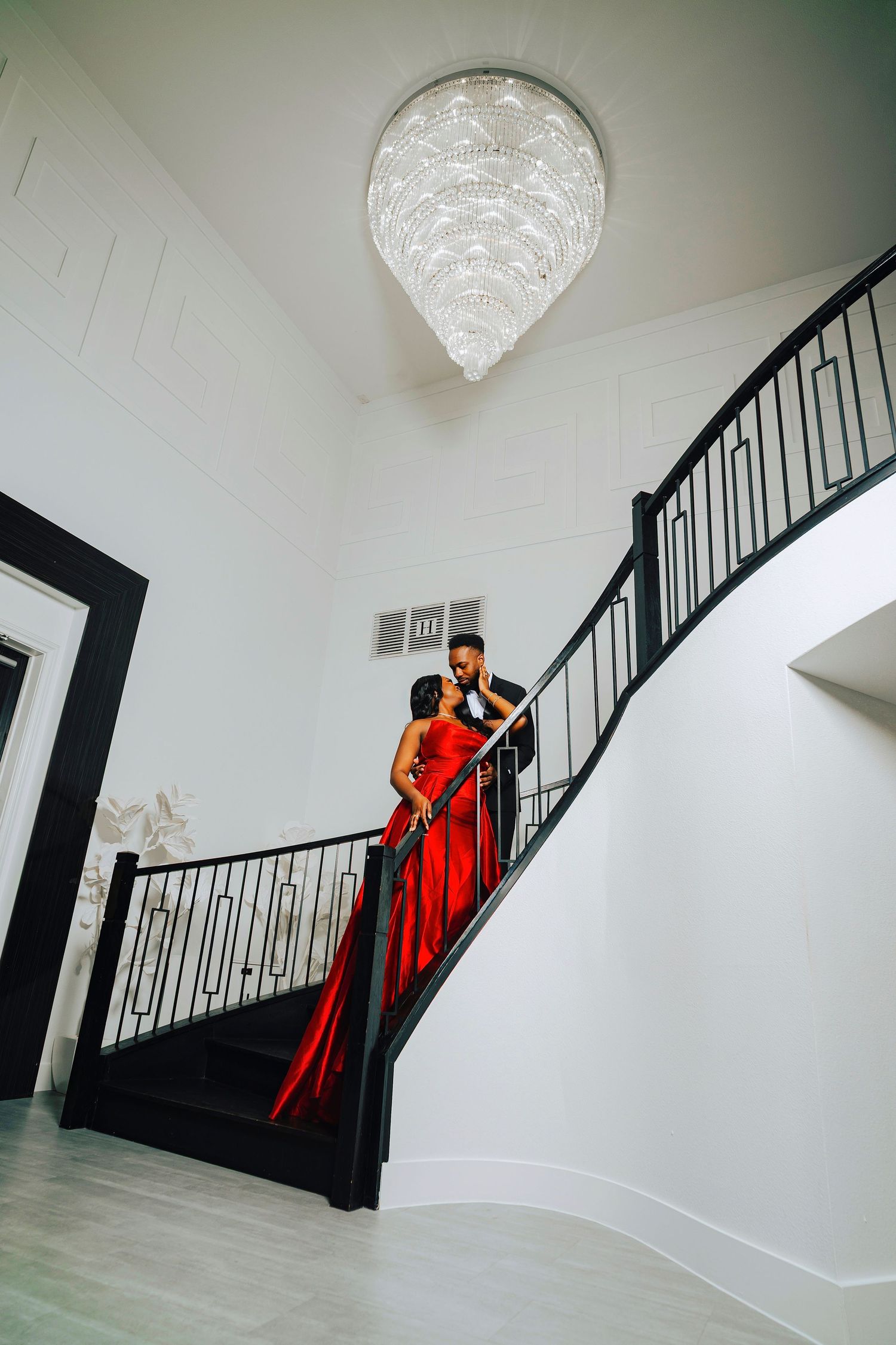 A figure in a flowing red dress poses dramatically on a staircase with black railings against white walls and crystal chandelier.
