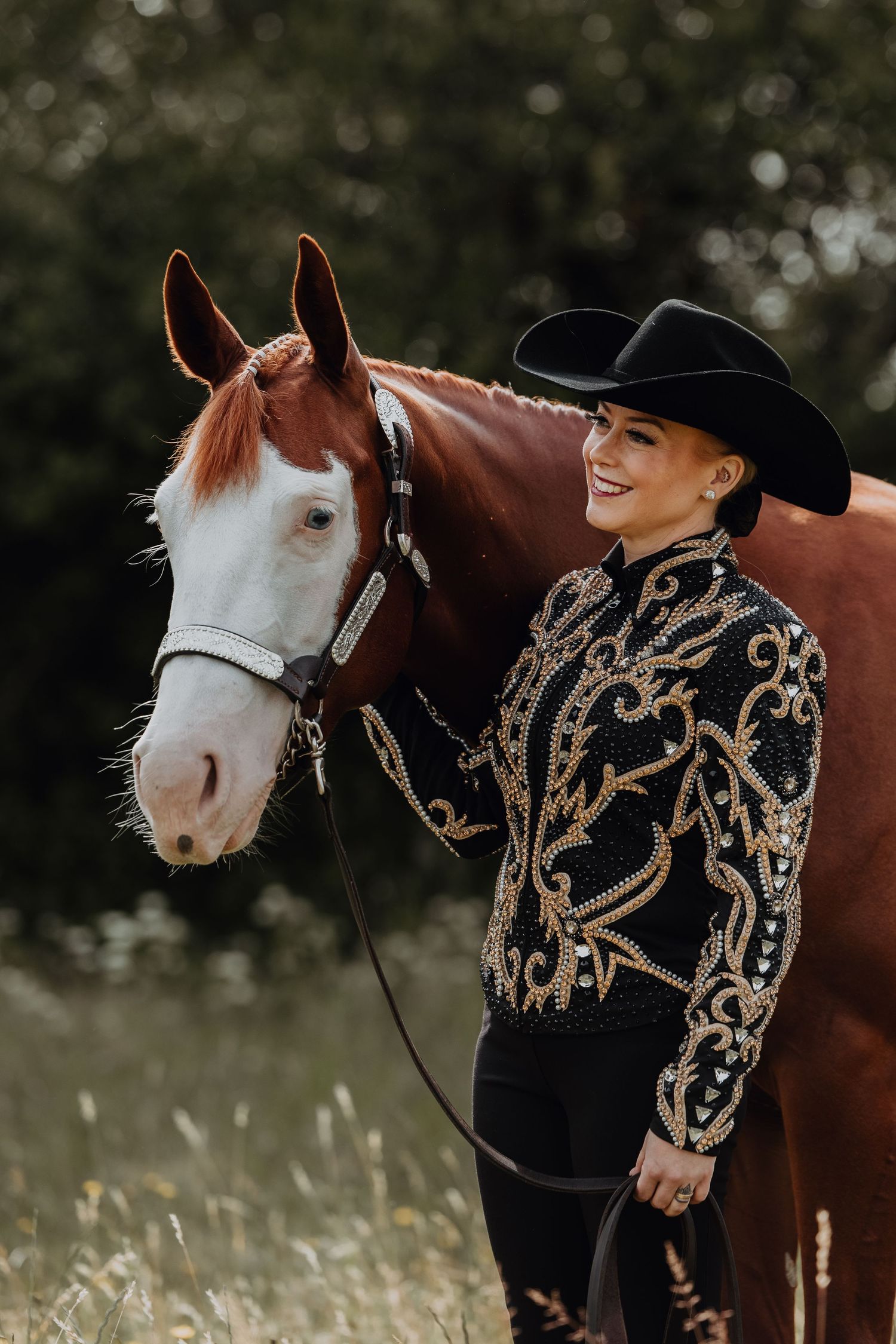 Horse with distinctive white blaze wearing bridle stands beside rider in ornate western show attire.