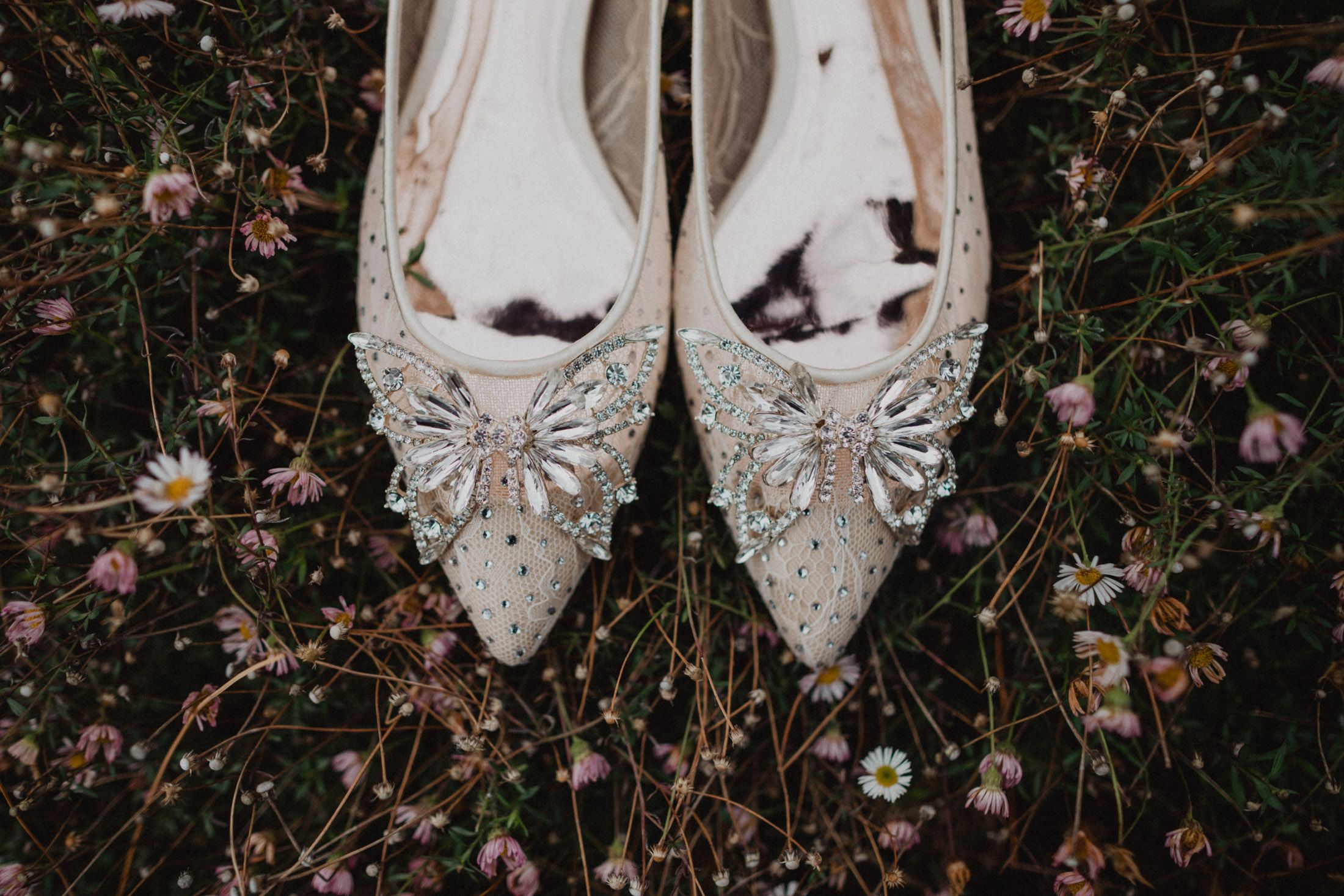 A pair of white leather ballet flats with floral embellishments rests on dark foliage and scattered pink petals.