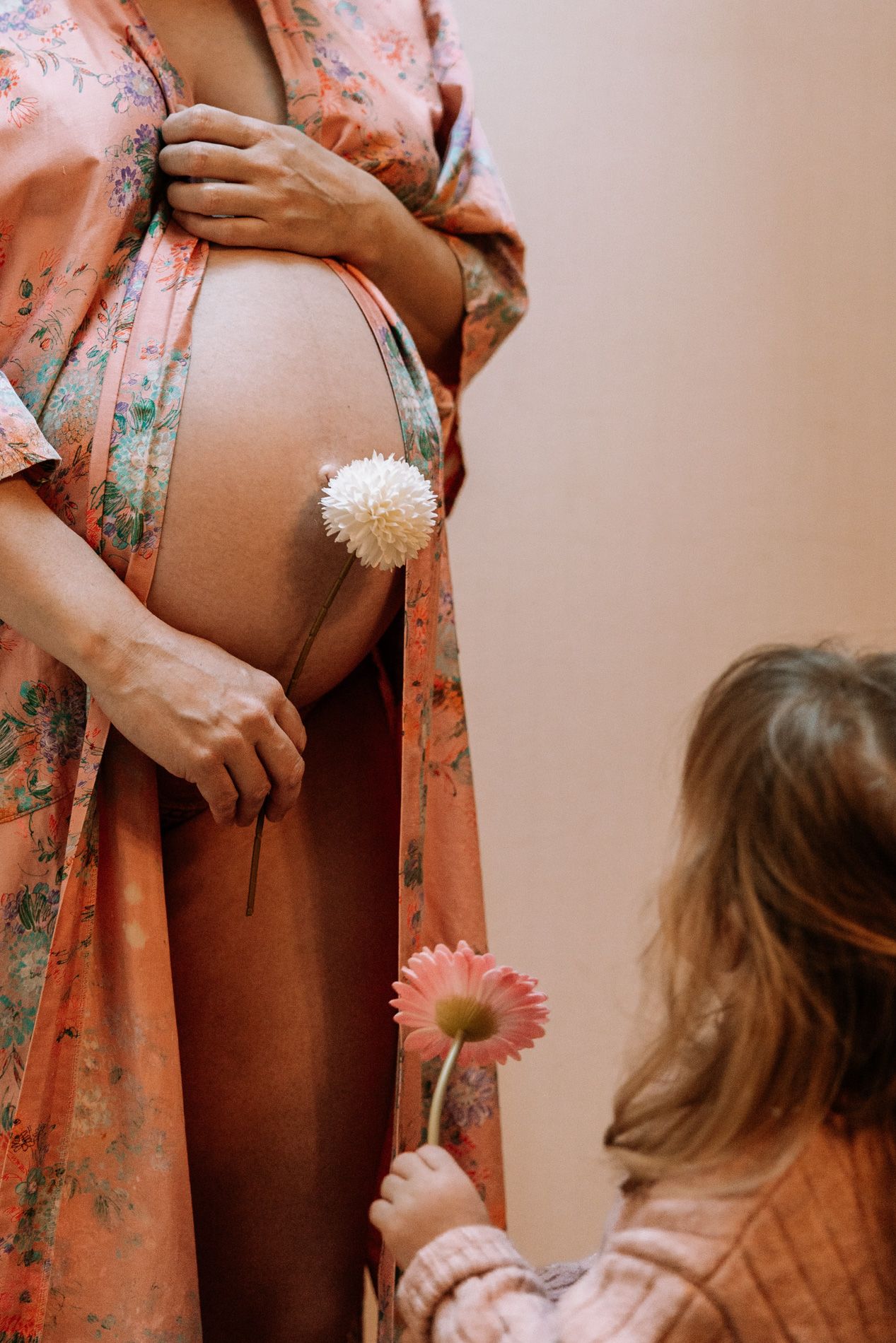 A pregnant woman in a floral robe holds her belly while flowers are held near her baby bump in a maternity photoshoot.
