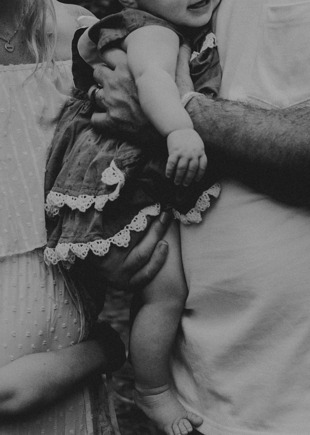 A vintage black and white photo showing a baby wearing a dress with delicate lace trim being held.
