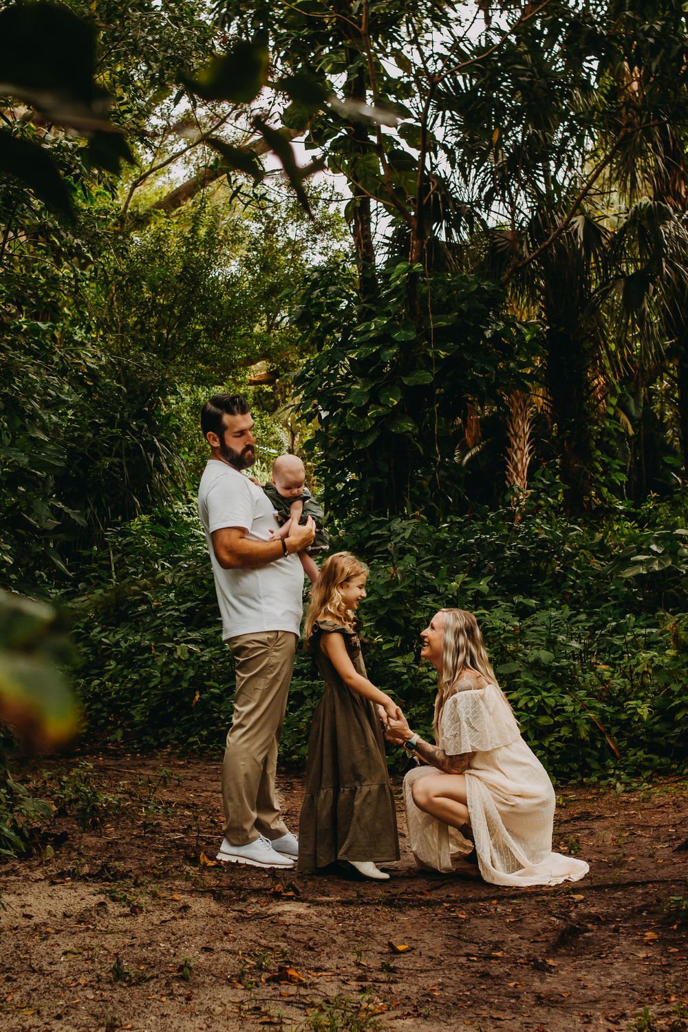 A family shares a tender moment together while kneeling on a dirt path surrounded by lush greenery in a forest setting.