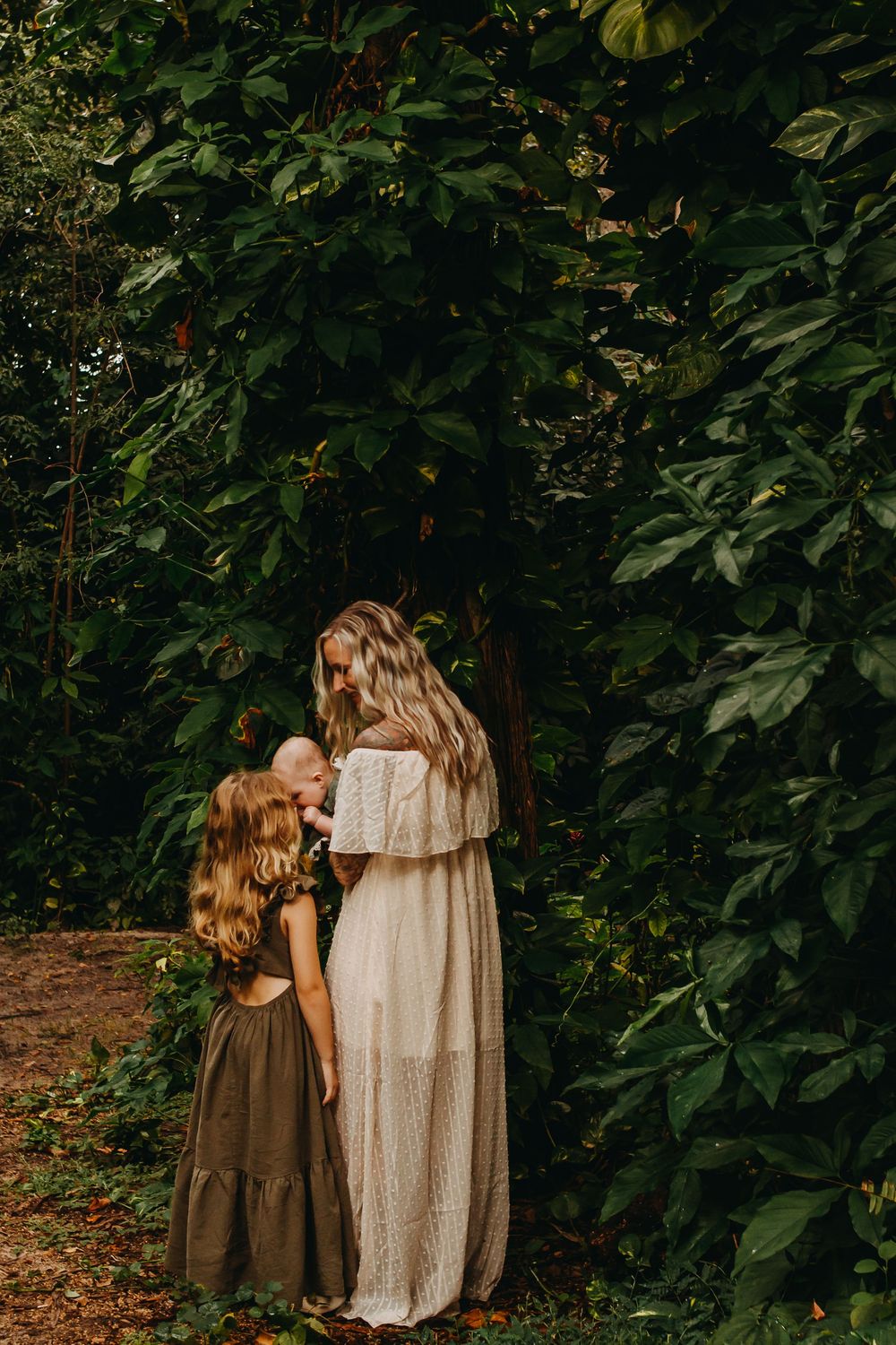 Two figures in flowing dresses embrace tenderly among dense foliage in a wooded outdoor setting.