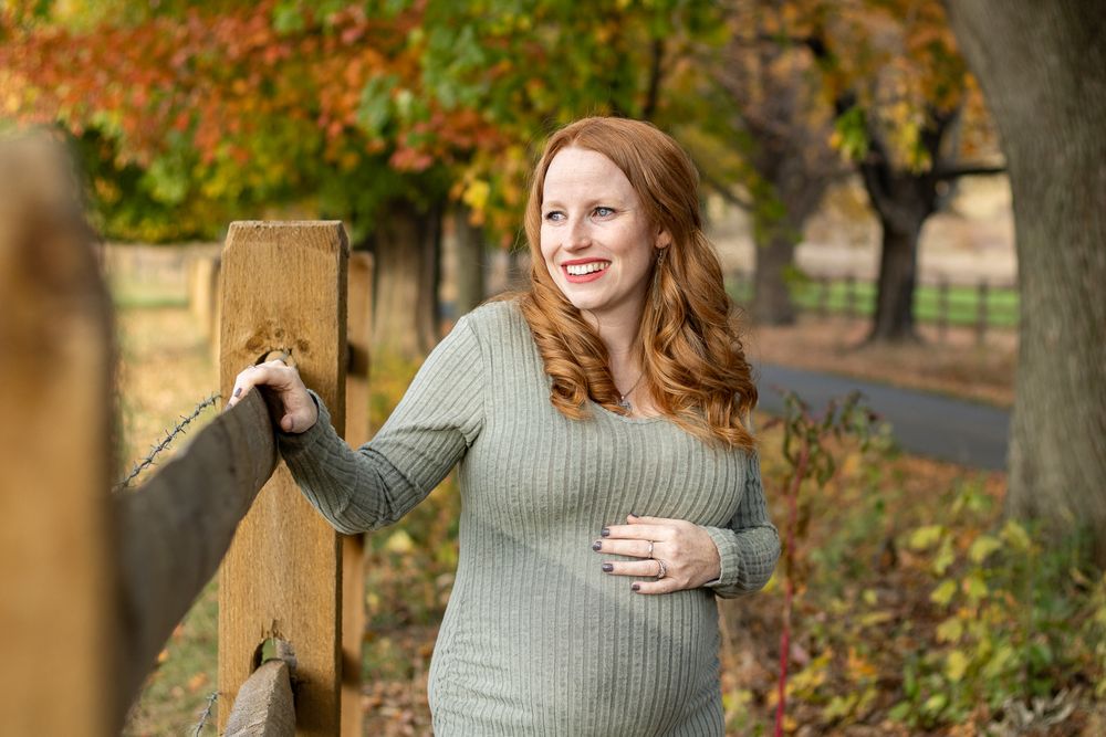 A red-headed maternal portrait of a woman in a pale green sweater who stands by a wooden fence in autumn with colorful fall foliage.
