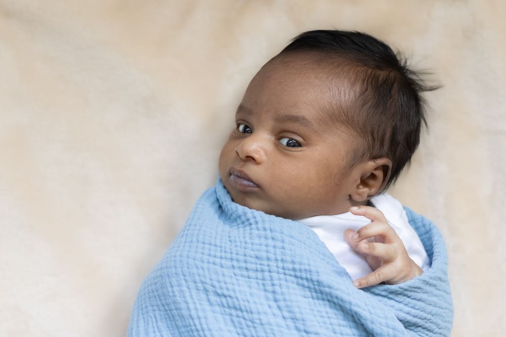 Newborn baby wrapped in soft blue blanket gazes upward while lying on beige surface, one hand escaped from the swaddle.