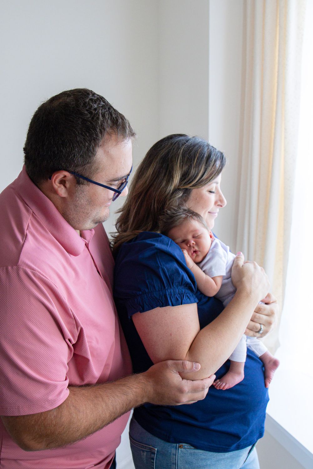 A couple in pink and navy-blue shirts share an intimate moment holding their newborn by a window.