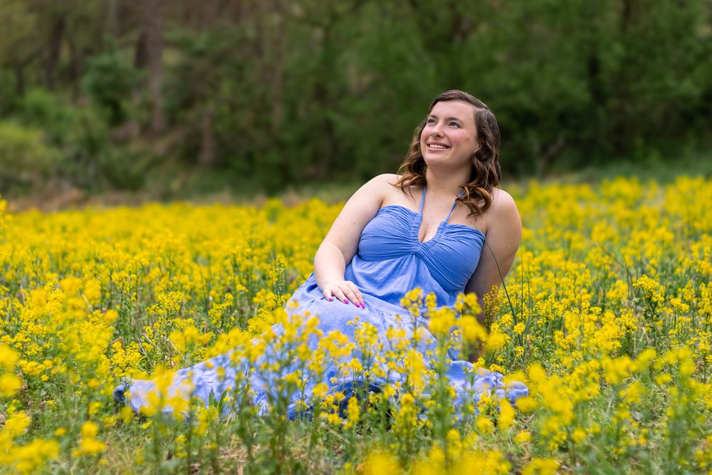 A high school senior in a blue dress sits among vibrant yellow wildflowers in a sunny meadow during springtime.