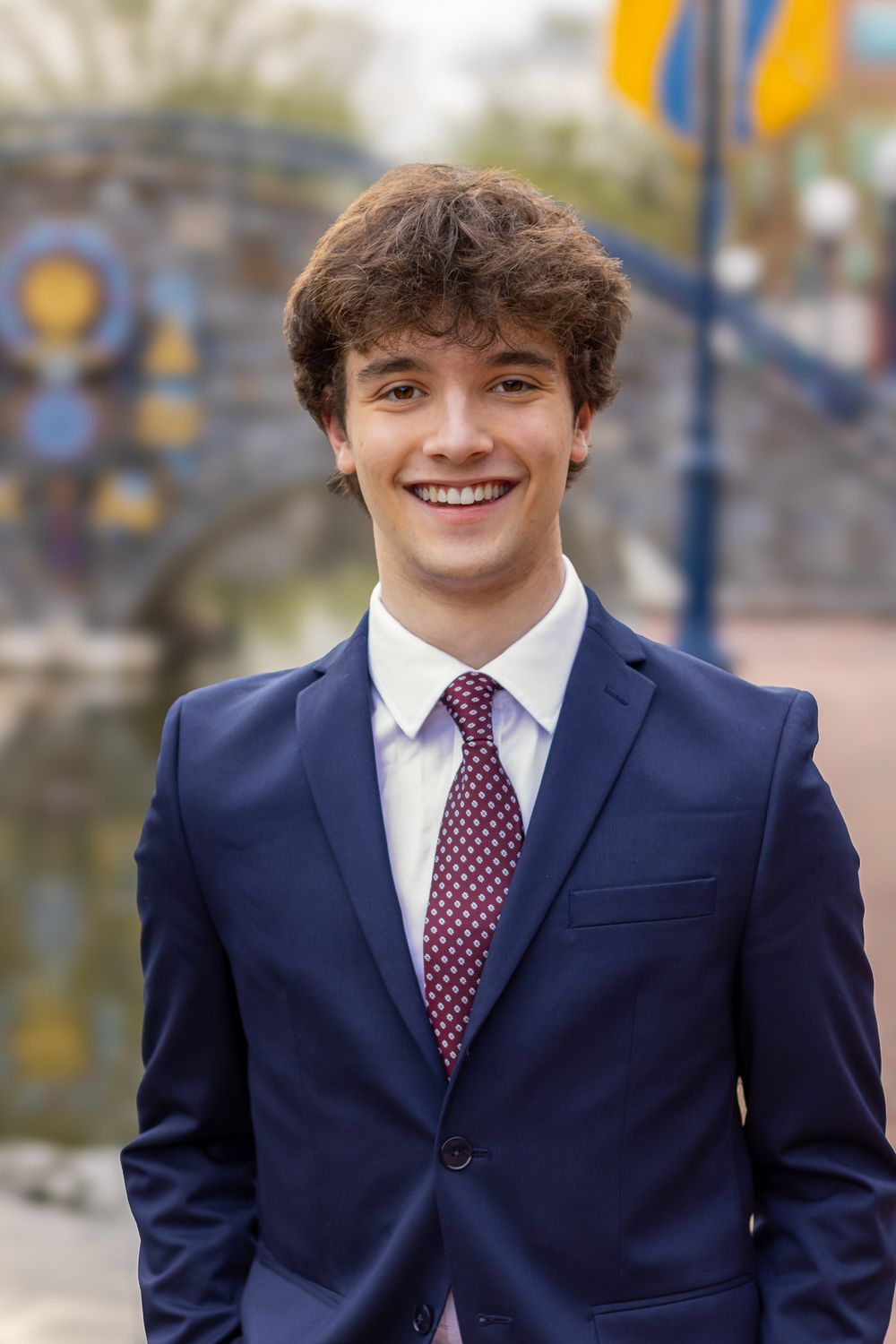 A high school senior in a navy-blue suit and burgundy polka dot tie poses outdoors for a formal portrait.