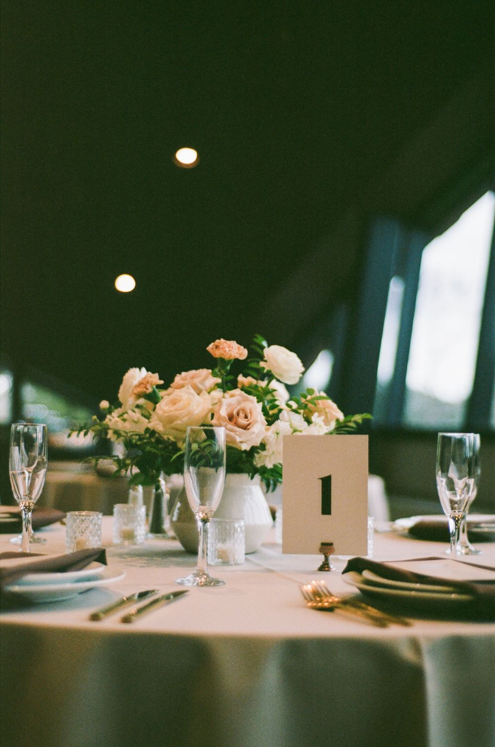 Elegant wedding reception table settings with soft pink floral centerpieces and crystal stemware in a dimly lit venue.