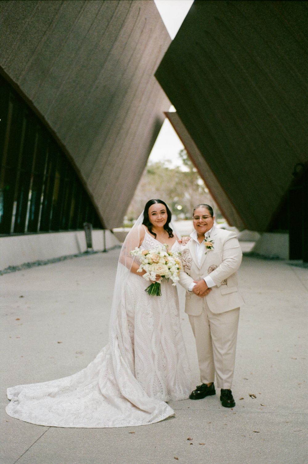 Two people in wedding attire pose together in front of an architectural structure with angular walls.