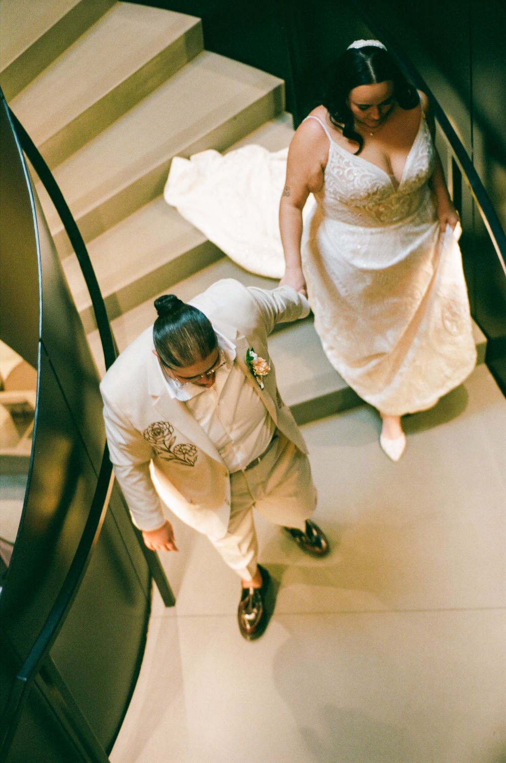 High angle view of a bride in a white wedding gown walking down a staircase showing the dress's flowing details.