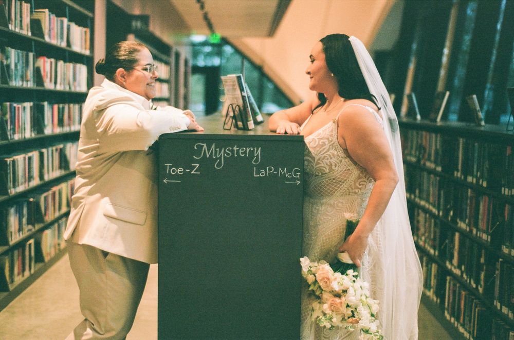 Two people pose playfully with a chalkboard sign reading 'Mystery' between library bookshelves.