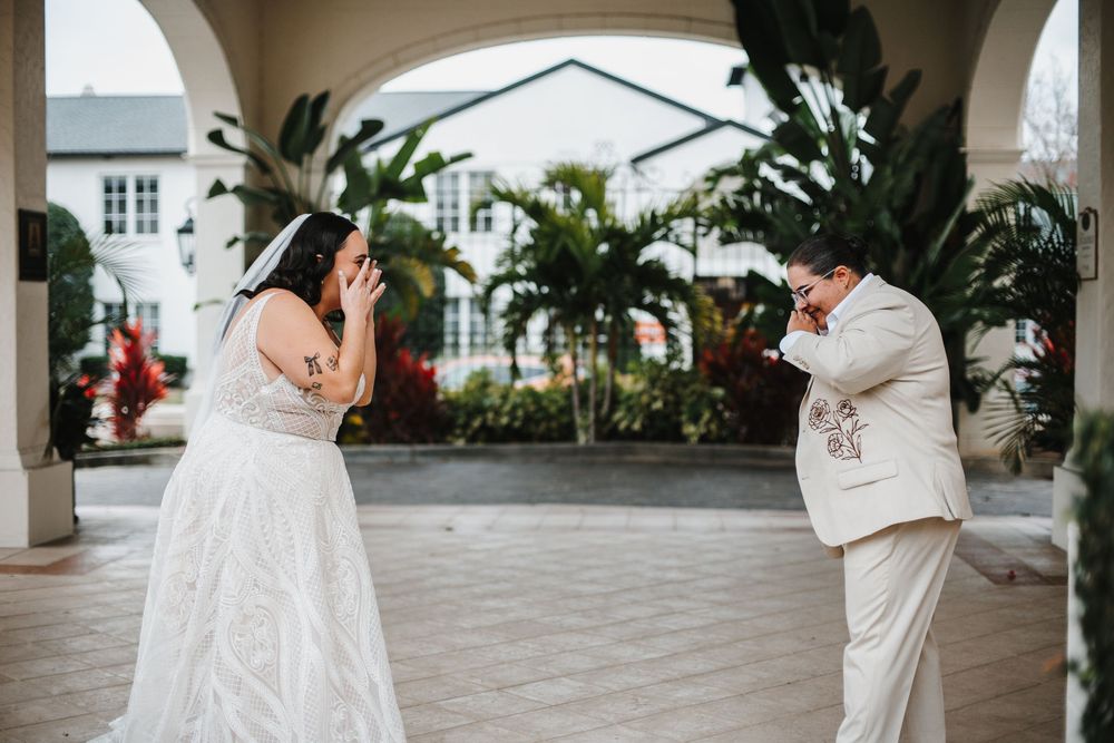 Two people in white attire share an emotional first look moment in a tropical courtyard setting.